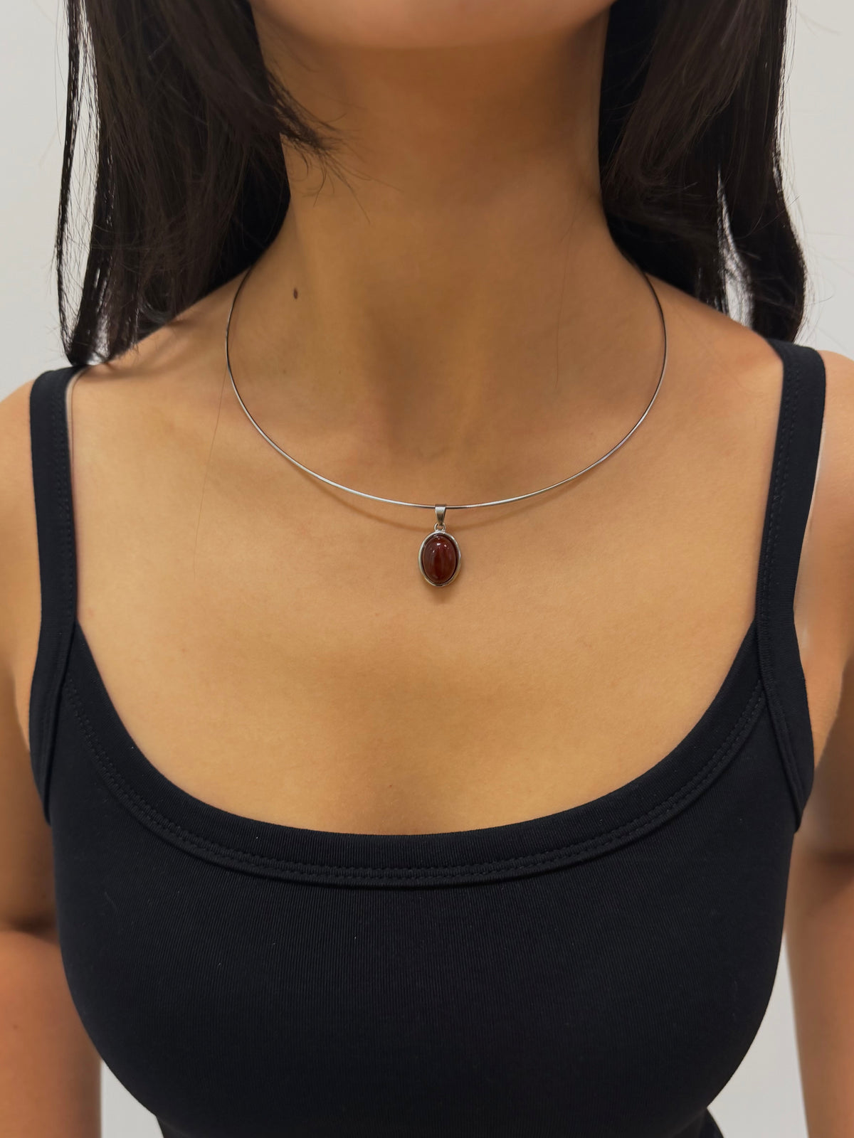 A close-up of a model wearing a delicate silver wire necklace with an oval dark red gemstone pendant, paired with a black tank top.