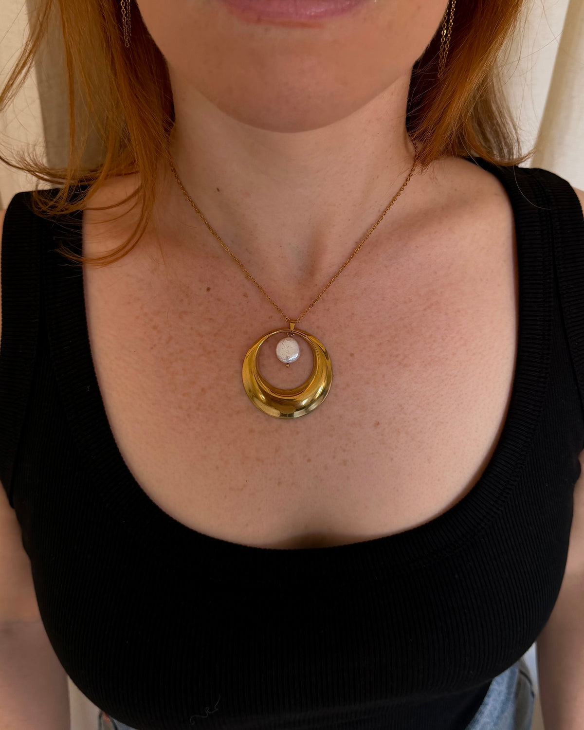 A close-up of a woman wearing a gold crescent moon pendant necklace with a white pearl center, styled with a black ribbed tank top and light blue denim jeans.