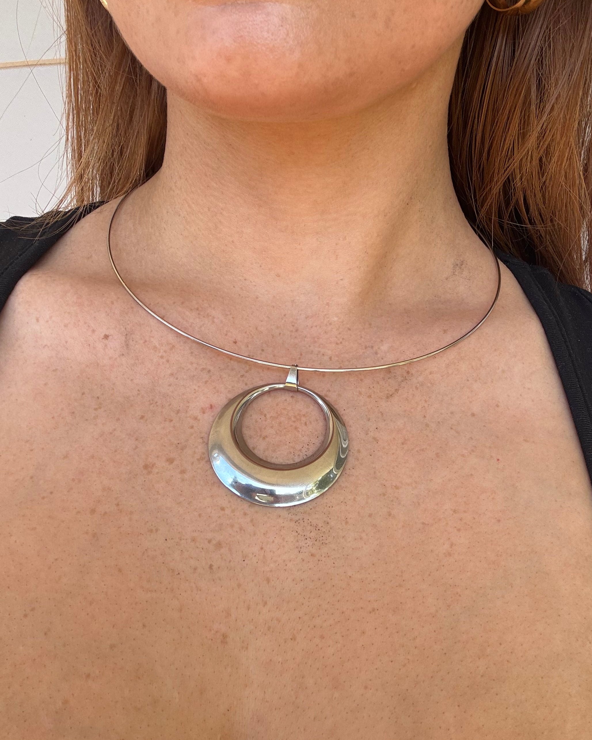 A close-up of a person's neck and upper chest wearing a silver-toned circular collar necklace with a thin wire loop extending from the base of the neck.