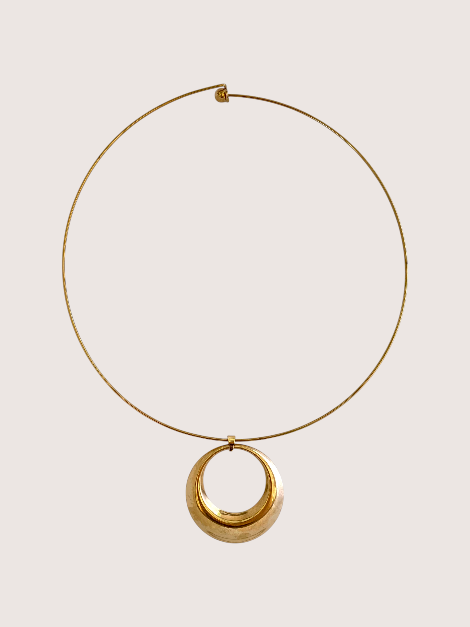 A gold-toned necklace featuring a thin circular wire hoop with a larger, polished concentric circular pendant, displayed against a solid light beige background.
