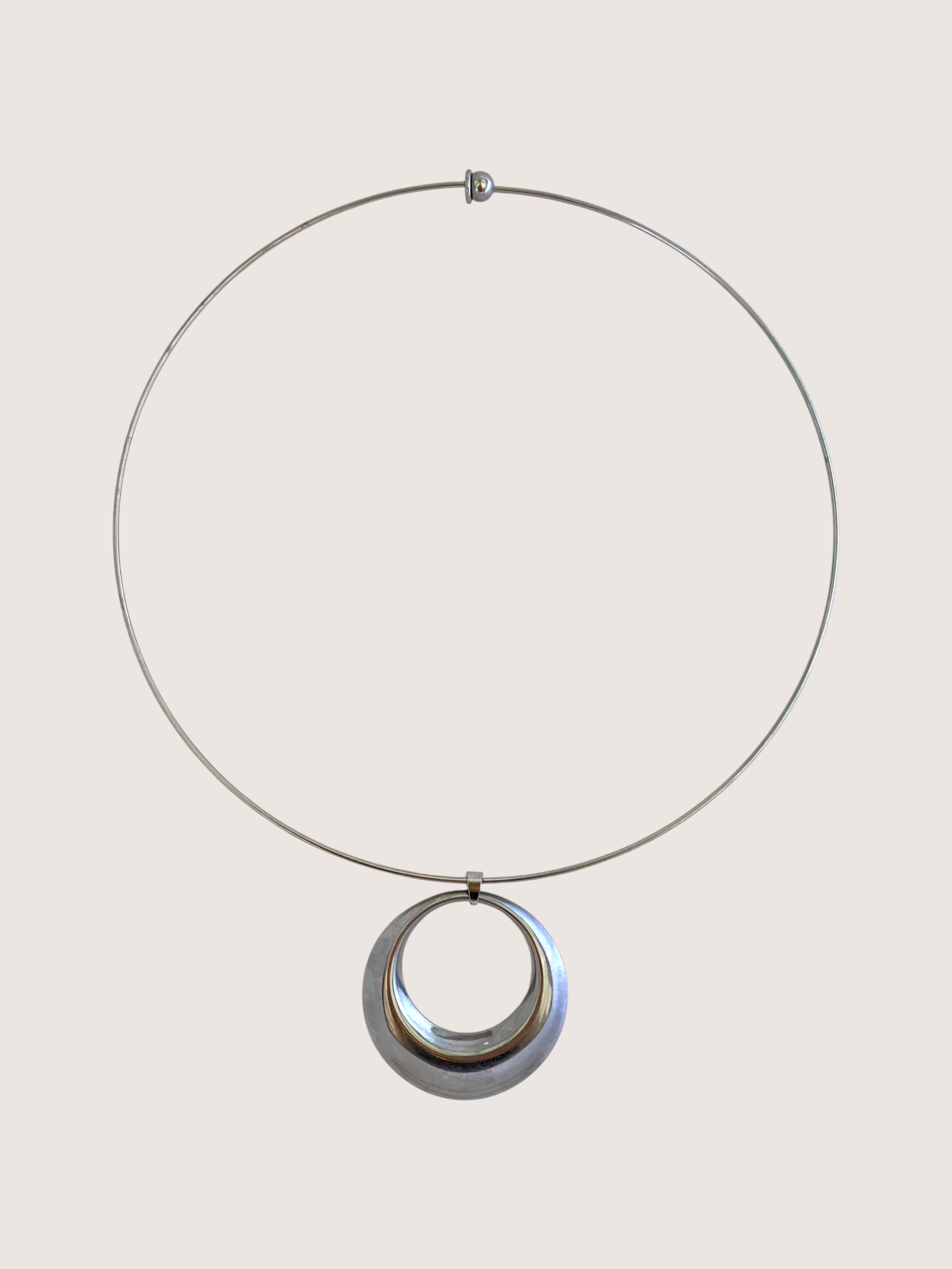 A minimalist silver-toned necklace featuring a thin circular wire hoop and a larger, polished concentric circular pendant, displayed against a solid light beige background.