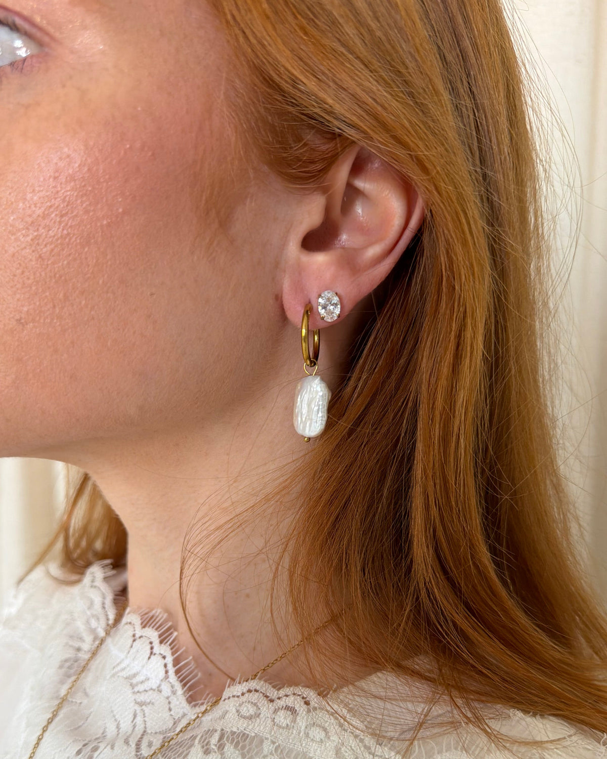 A close-up of a woman wearing a gold huggie hoop earring with a white textured drop pendant and a clear crystal stud, paired with a white lace top.