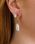 A close-up of a person's ear wearing a gold huggie hoop earring with a dangling baroque pearl and a small clear crystal stud.