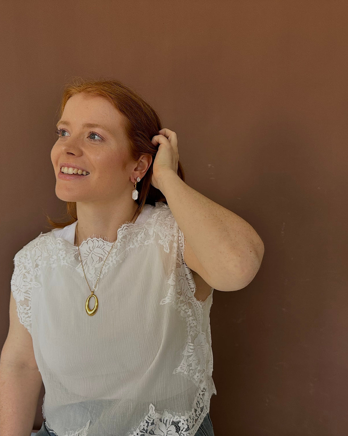A smiling woman with red hair looking upward and touching her hair, wearing a white lace-trimmed blouse and a gold pendant necklace, standing against a solid brown wall.