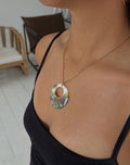 A close-up lifestyle shot of a model wearing a gold chain necklace with a circular abalone shell pendant. The pendant features a natural iridescent pattern with shades of green, blue, and pink.