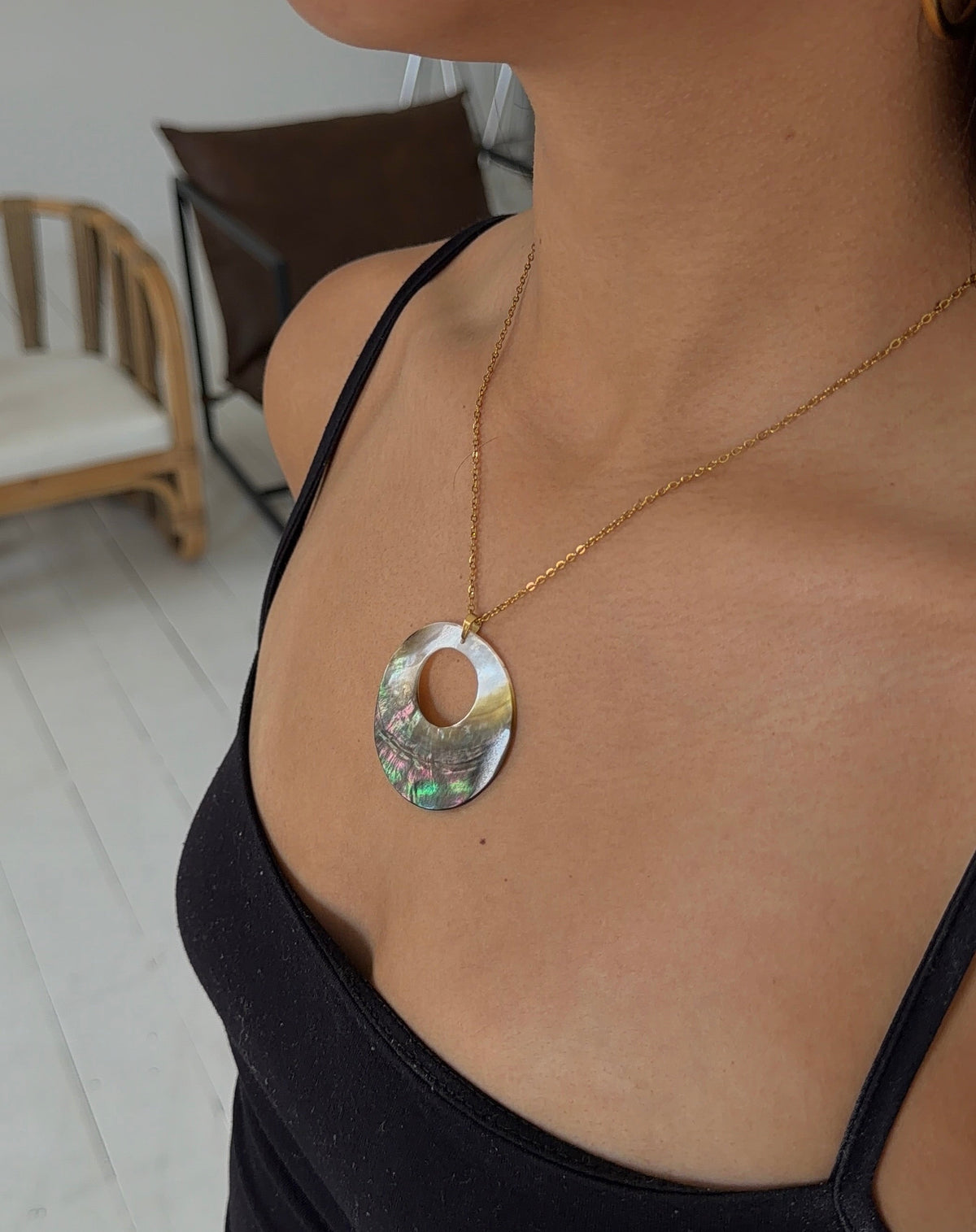 A close-up lifestyle shot of a model wearing a gold chain necklace with a circular abalone shell pendant. The pendant features a natural iridescent pattern with shades of green, blue, and pink.