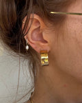 A close-up of a person's ear wearing a chunky, textured gold huggie hoop earring and a small silver hoop with a white pearl drop earring.