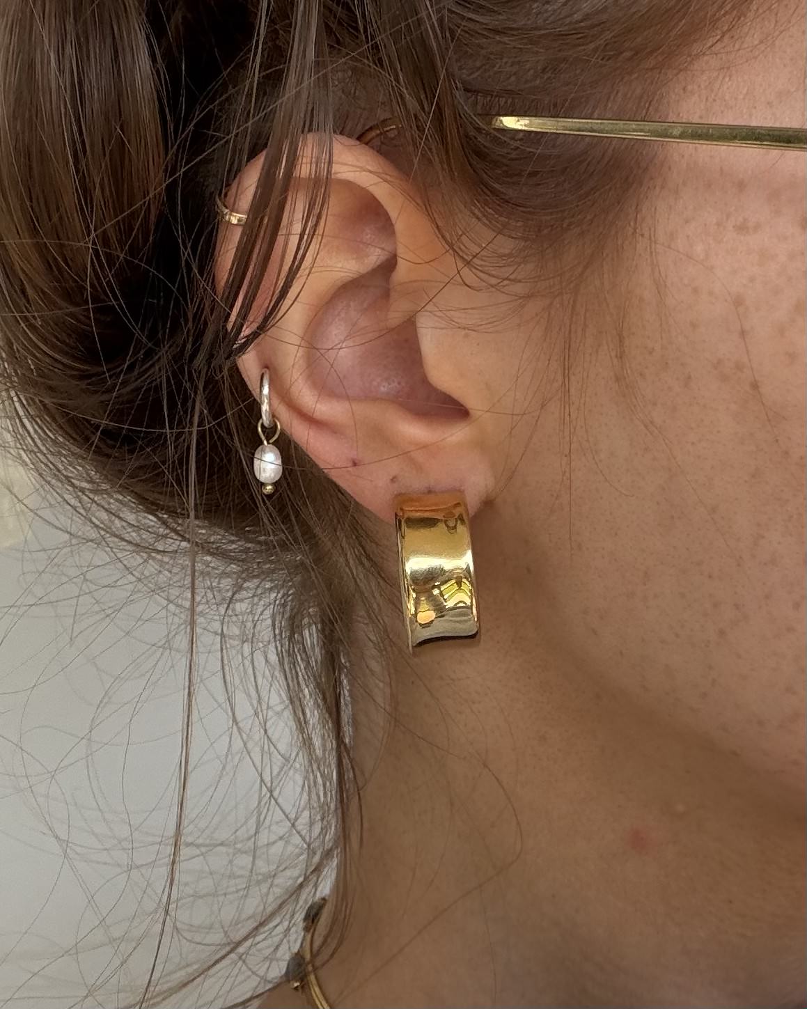 A close-up of a person's ear wearing a chunky, textured gold huggie hoop earring and a small silver hoop with a white pearl drop earring.