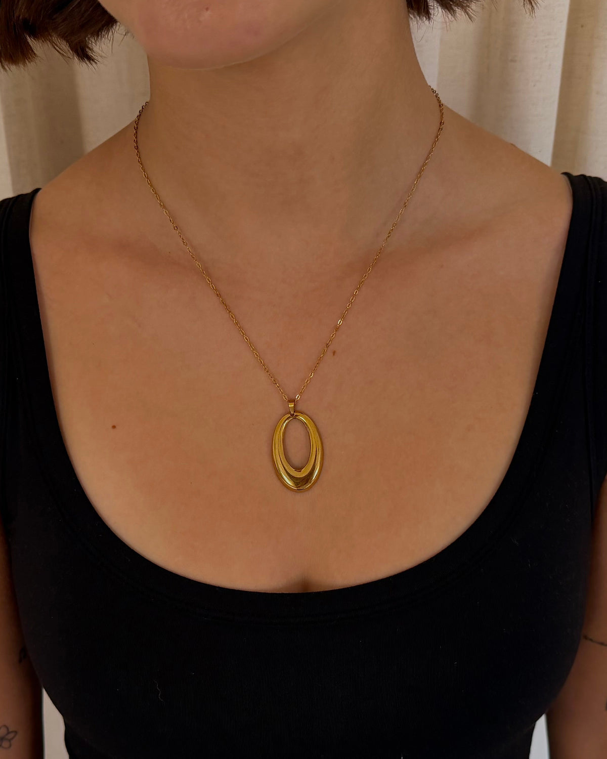 A person wearing a gold double-ring pendant necklace over a black tank top, standing in front of beige curtains.
