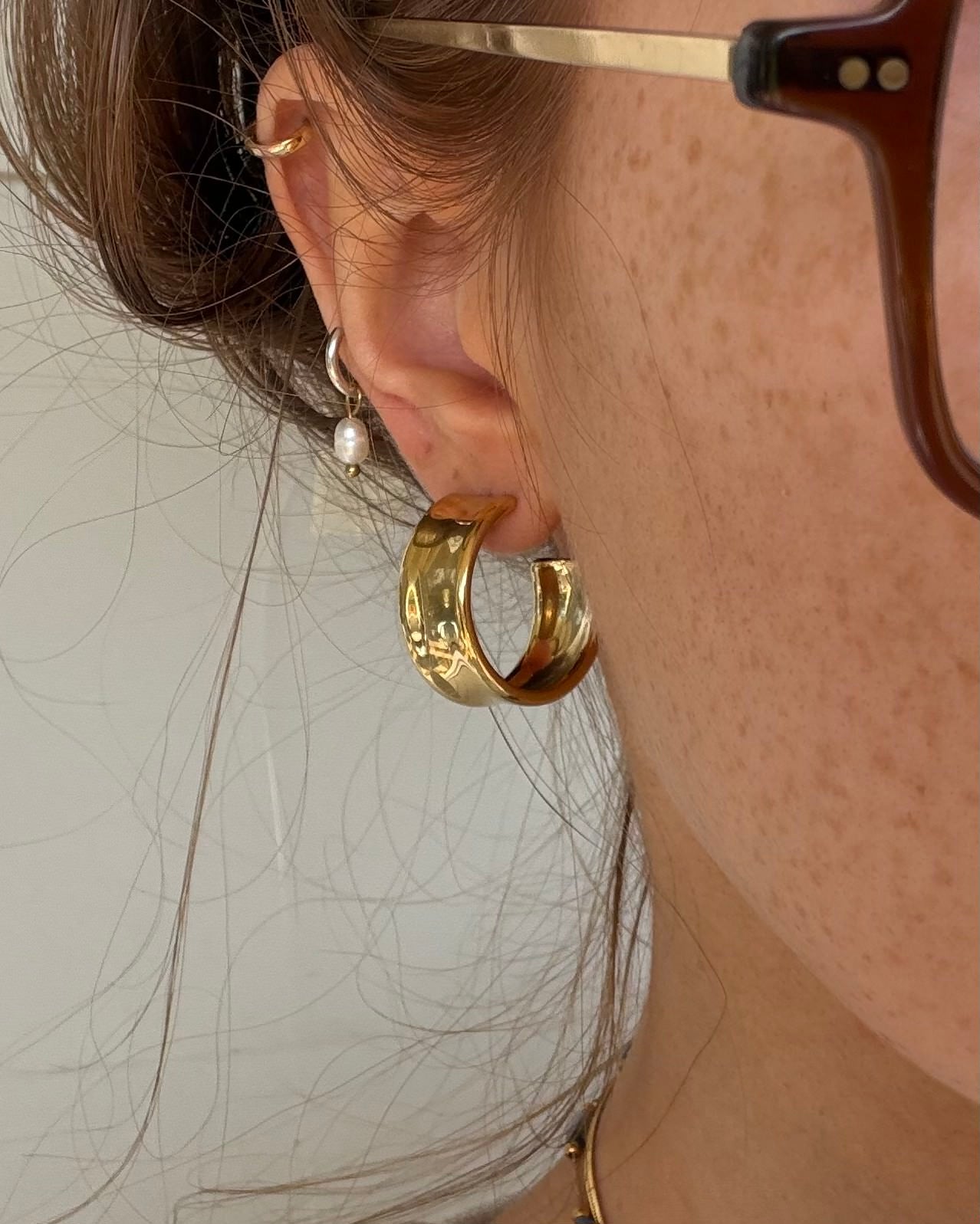 A close-up of a person's ear wearing a thick, polished gold hoop earring, along with other small gold and pearl earrings, brown hair, and part of a tortoiseshell eyeglass frame.