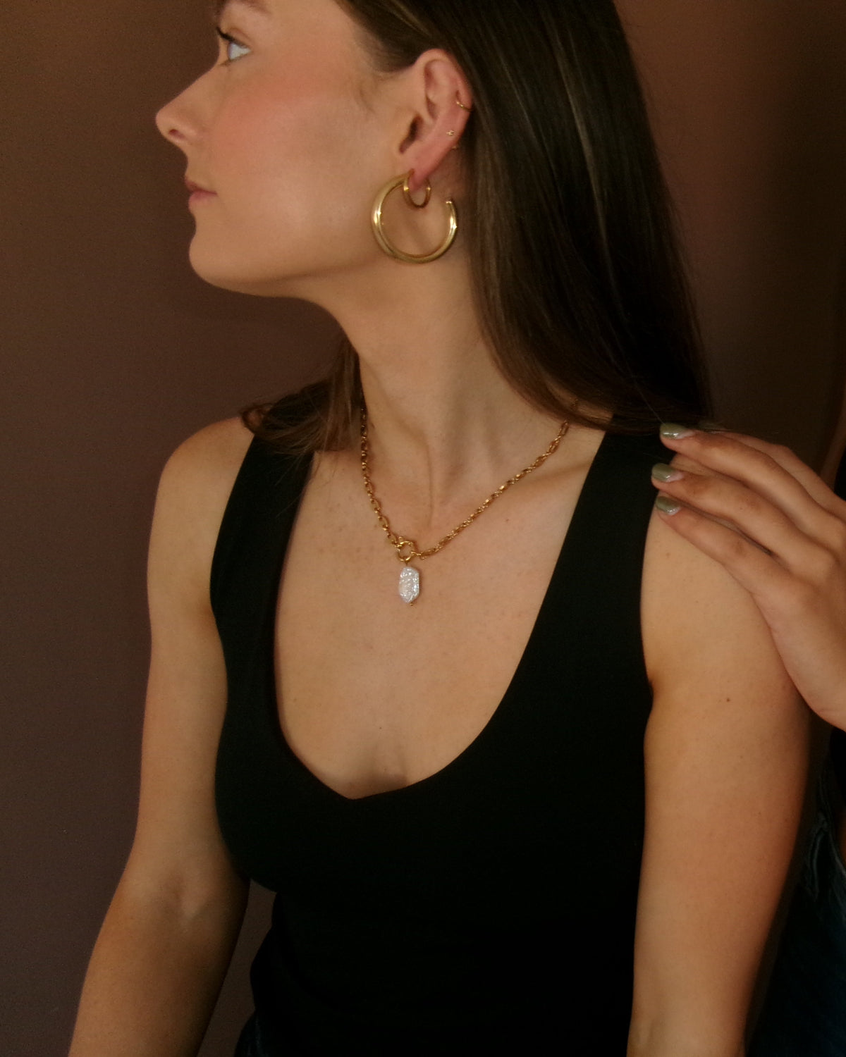Two women wearing black camisole tops and jewelry, with one placing a hand on the other's shoulder. Visible jewelry includes a gold hoop earring, a gold pendant necklace, and a turquoise beaded necklace.
