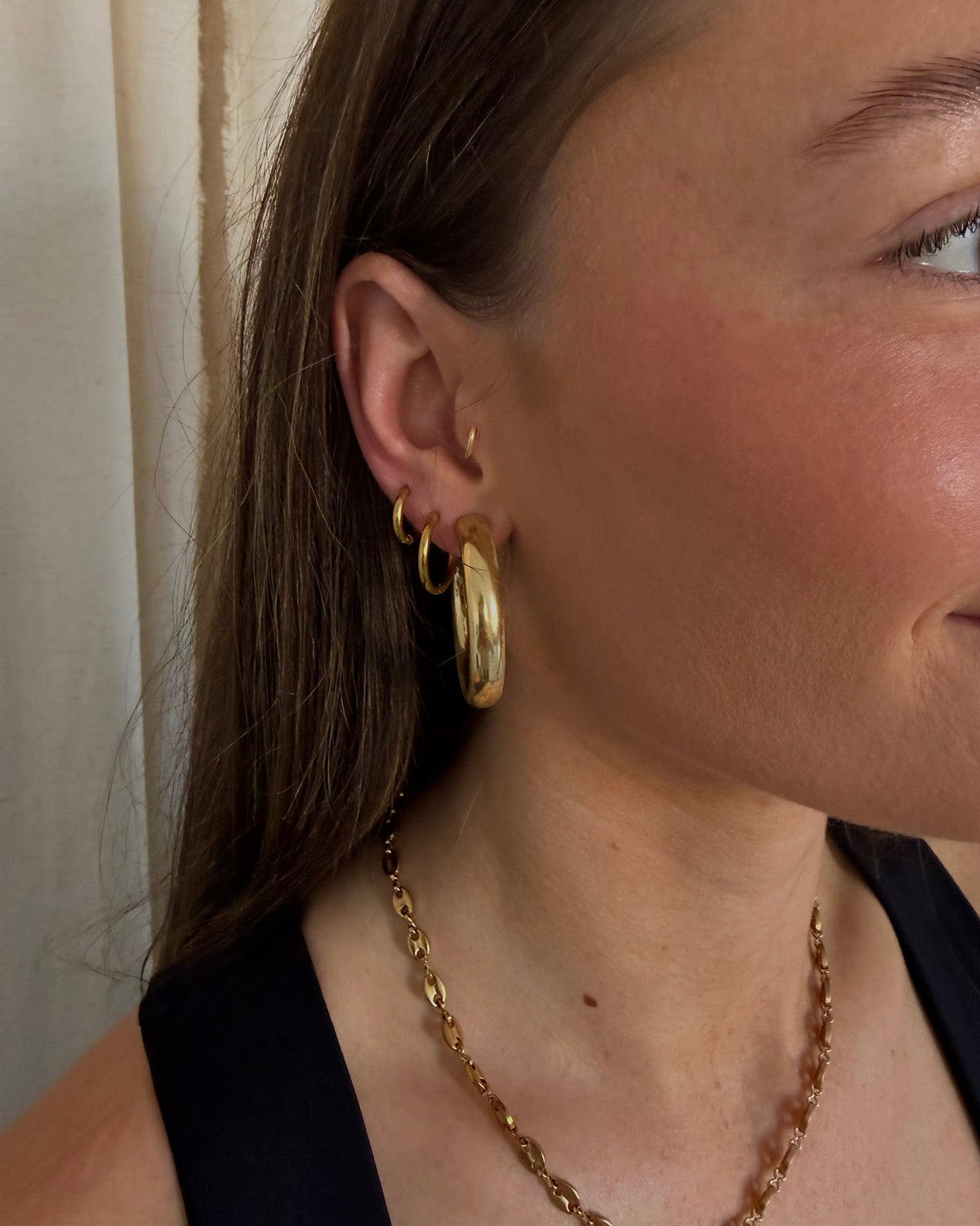 A close-up of a woman's ear wearing a stack of gold hoop earrings, including one large textured gold hoop and two smaller gold rings, along with a gold chain necklace.
