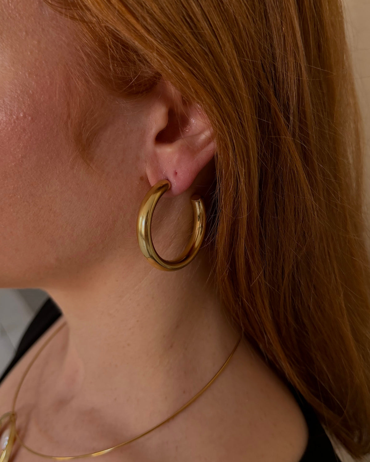A close-up side profile of a woman with red hair wearing a thick gold hoop earring and a delicate gold necklace with a circular pendant, set against a soft beige background.