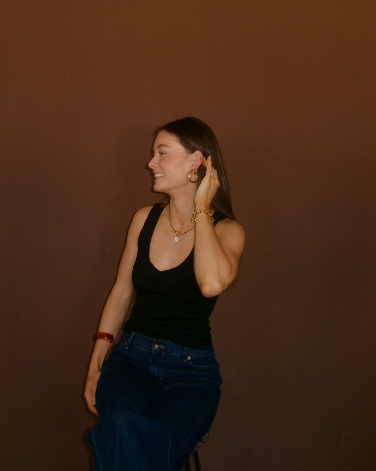 A woman wearing a black tank top and blue jeans stands against a solid dark brown background, smiling and looking to the side.