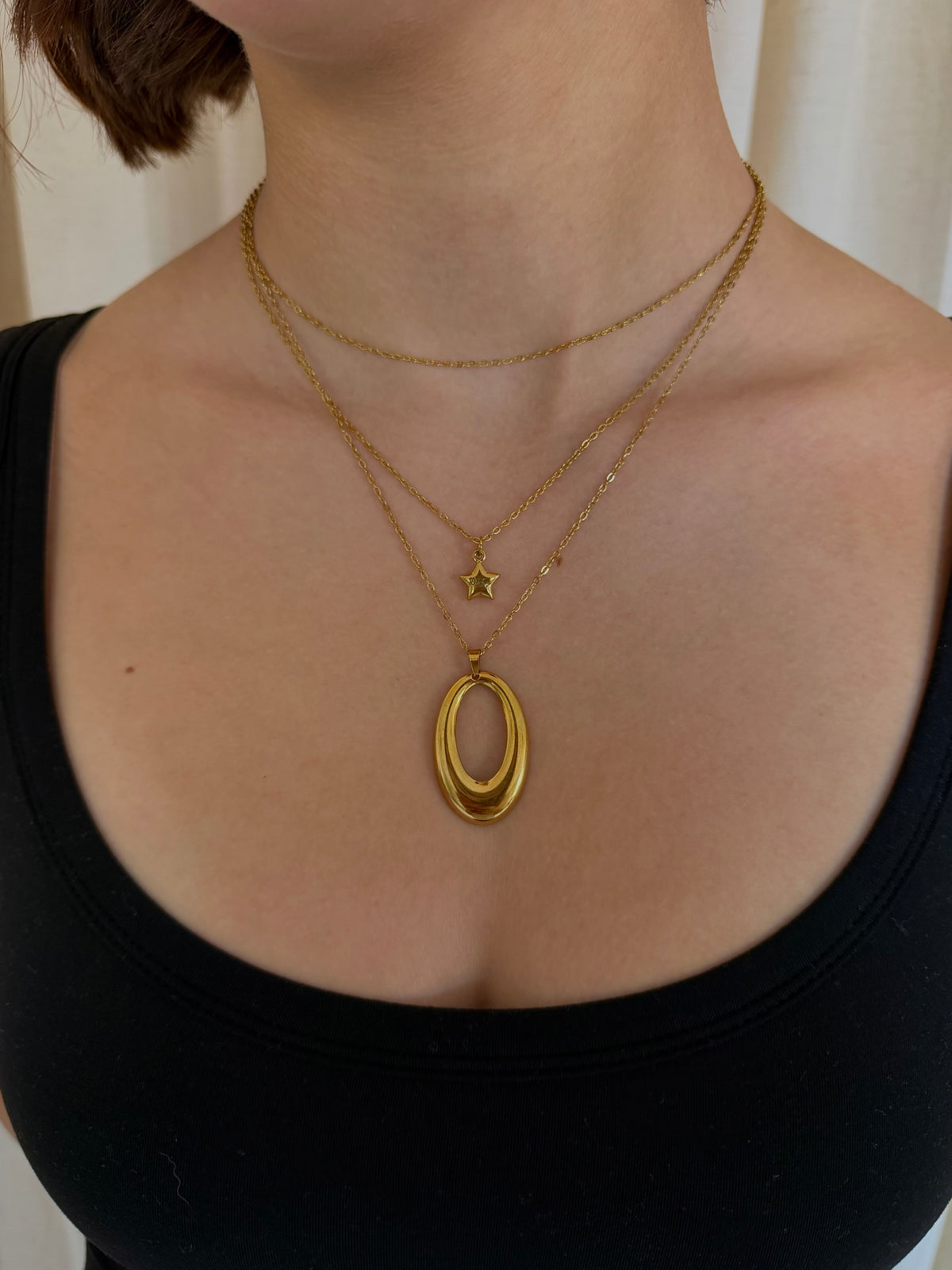 A close-up of a person wearing layered gold necklaces, including a large textured oval pendant, a small star charm, and a delicate chain, paired with a black top.