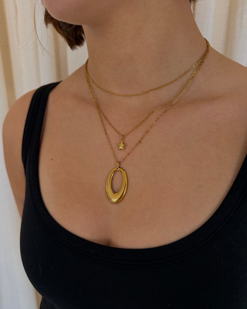 A close-up of a model wearing a layered gold necklace set, featuring a large polished oval pendant on a longer chain, a small star charm on a shorter chain, and a delicate chain layer, styled with a black tank top.