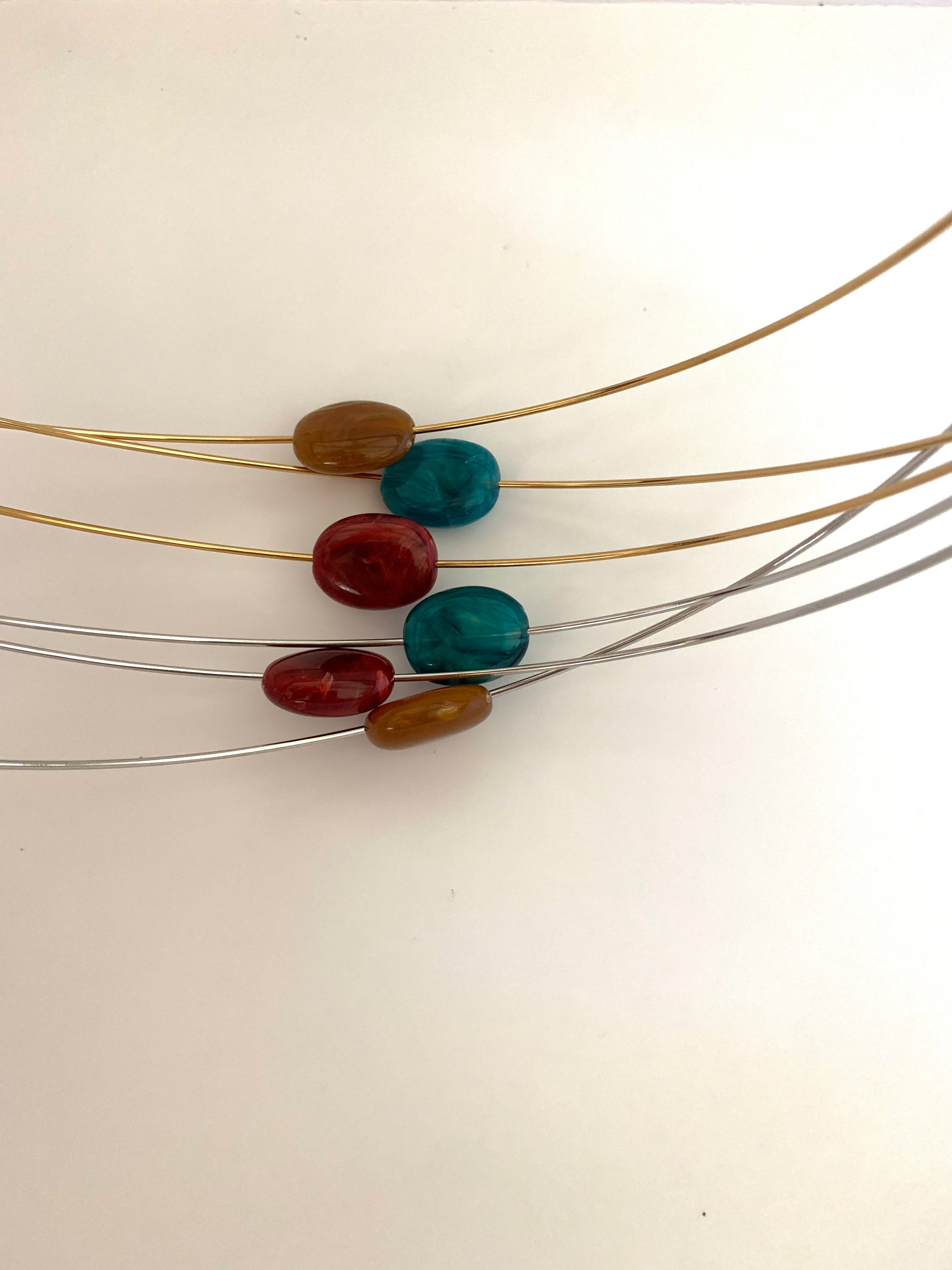A close-up of a jewelry-making component showing five polished cabochon beads in brown, teal, and red, threaded onto thin gold, silver, and gray metal wires against a plain off-white background.