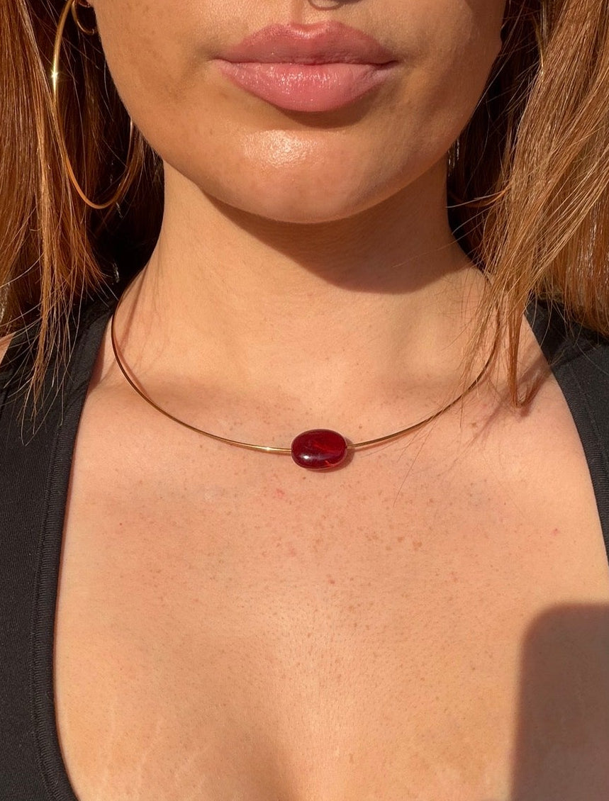 A close-up of a woman wearing a delicate gold chain necklace with a small oval dark red gemstone pendant, paired with a black sports bra.