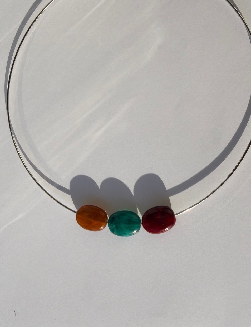 A thin metal circular necklace with three smooth, polished oval beads in amber, green, and dark red, displayed on a light gray surface.