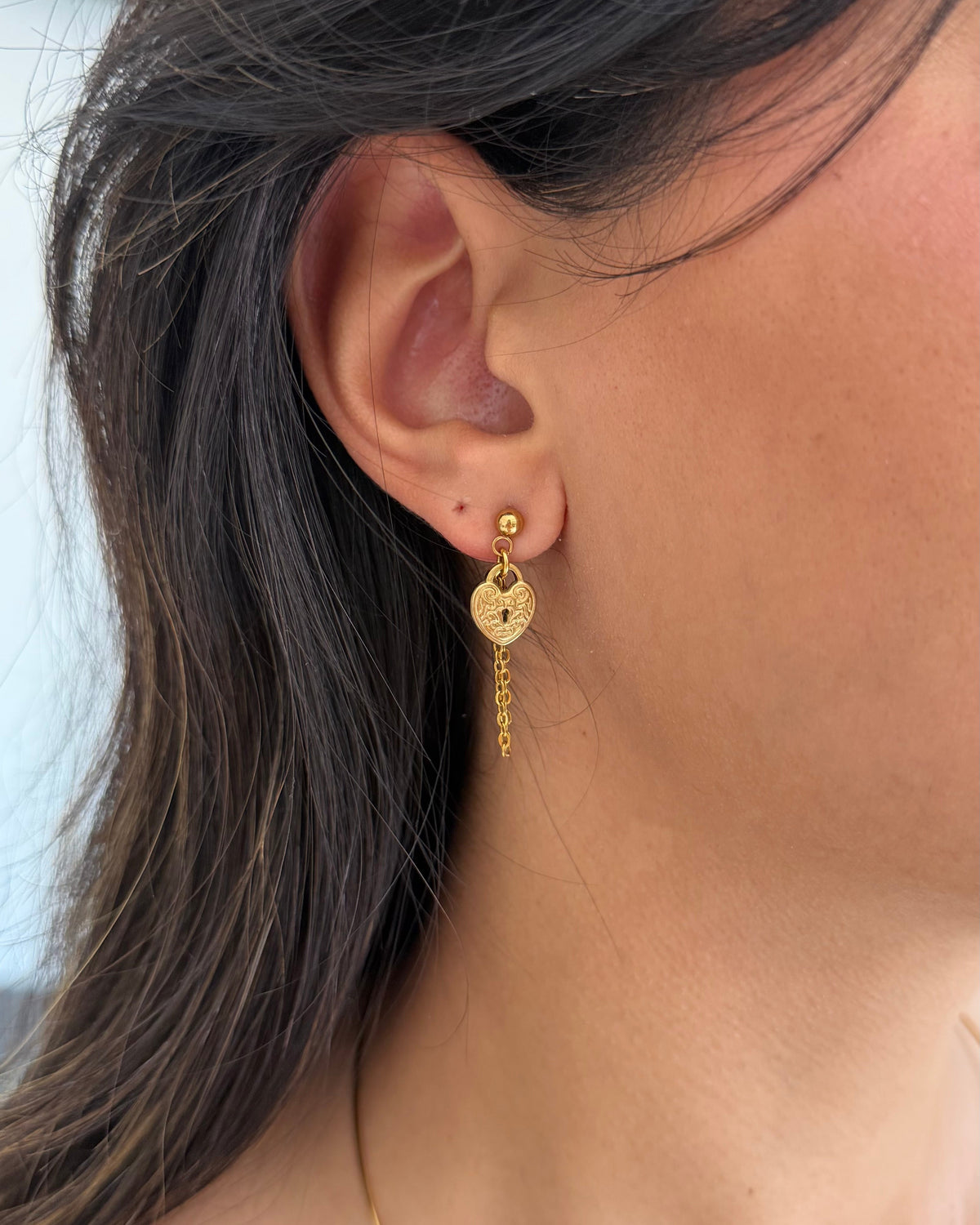A close-up of a person's ear wearing a gold dangle earring with a heart-shaped charm and a dangling chain.