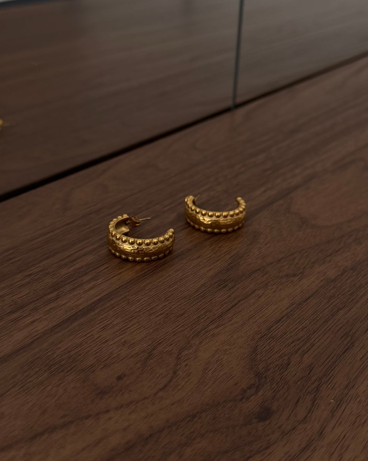A pair of textured gold crescent-shaped hoop earrings resting on a dark wood tabletop, with a glass partition and office chair visible in the background.
