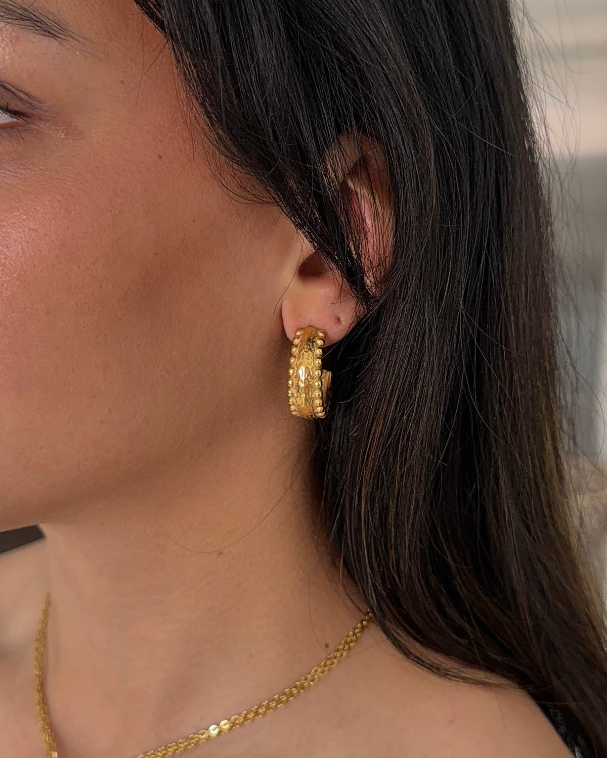 A close-up side profile of a woman wearing a gold hoop earring and a gold pendant necklace with a blue rectangular accent, against an indoor background.