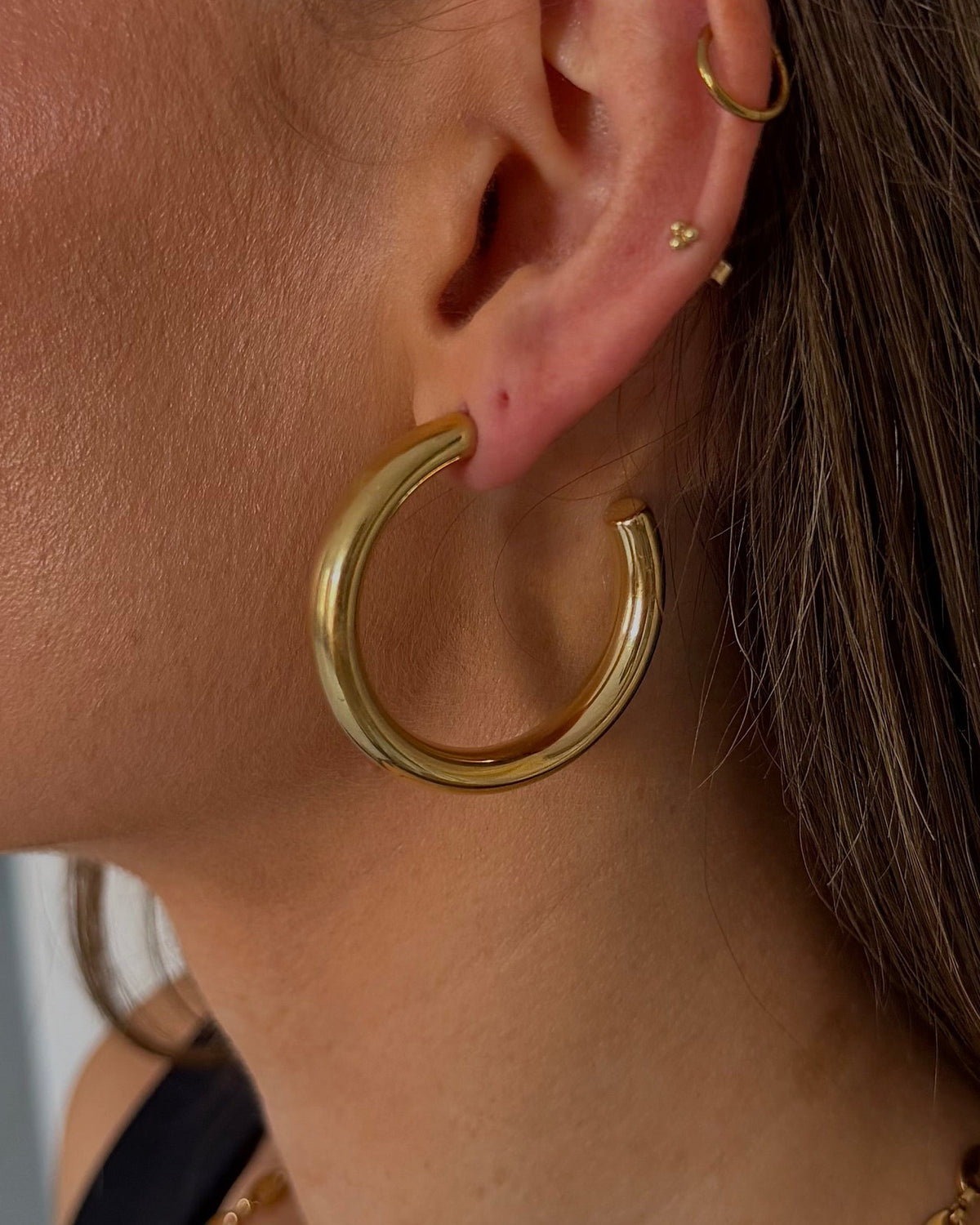 A close-up side profile of a woman wearing a thick, polished gold hoop earring, along with a gold chain necklace and small gold ear piercings.