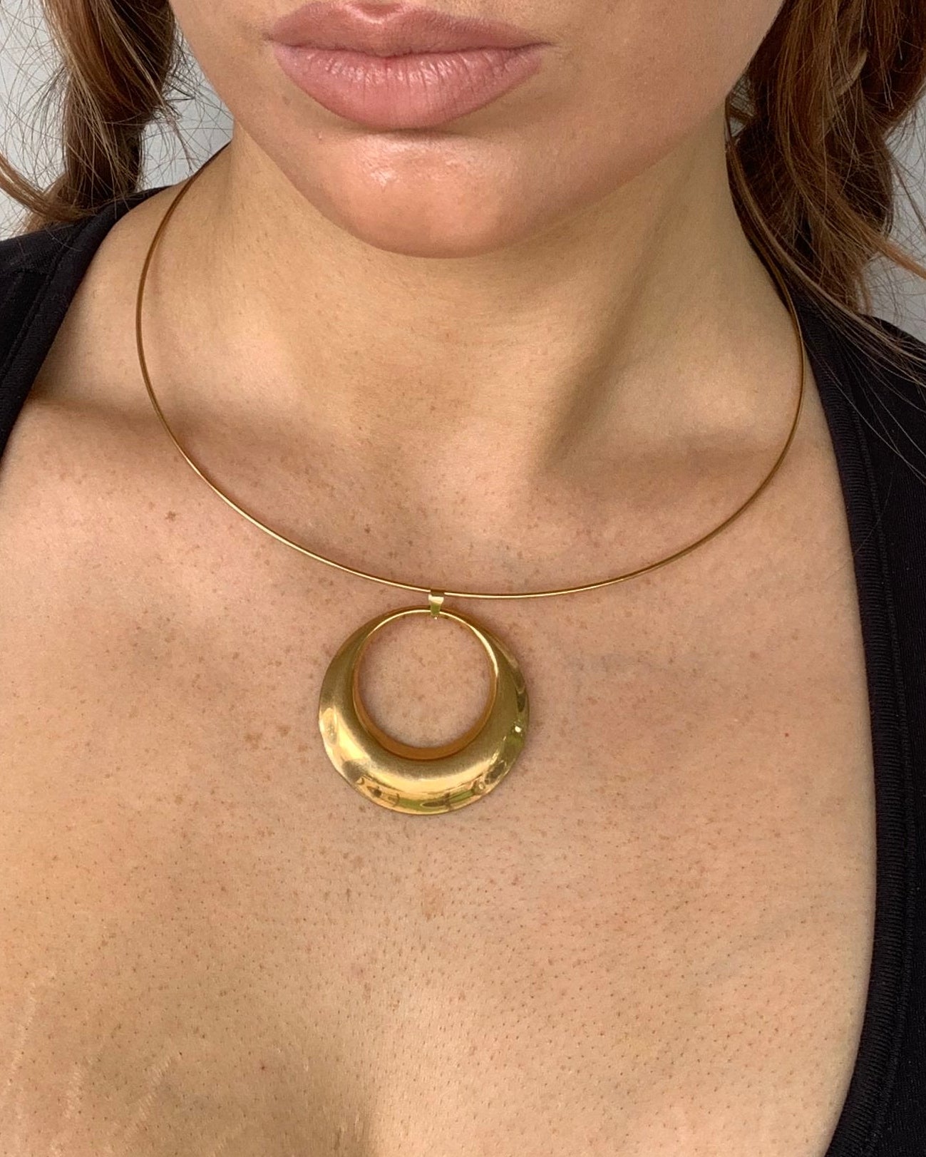 A close-up of a model's chest and neck area wearing a gold ring pendant necklace with a thin gold wire loop extending from the bottom of the ring.