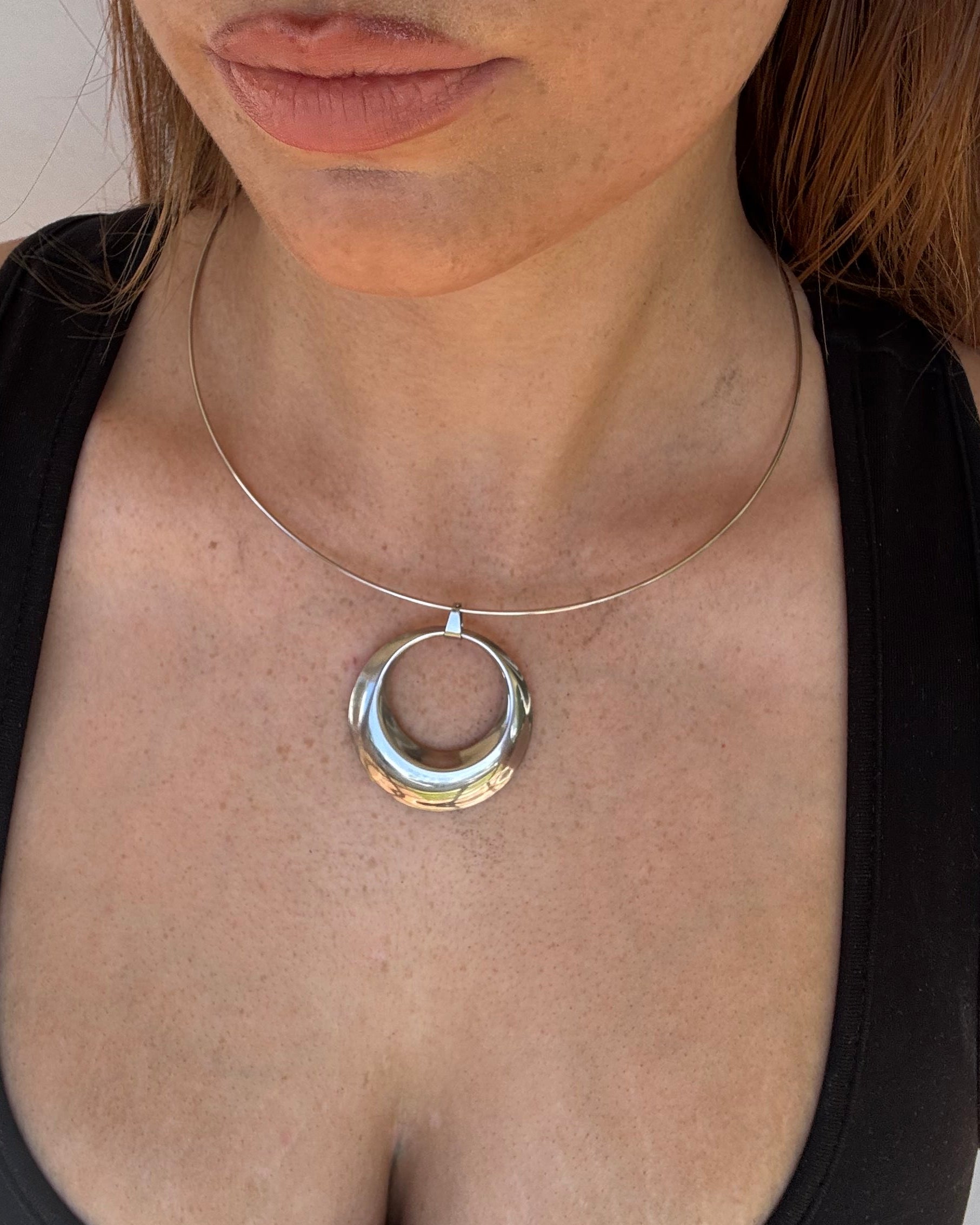 A close-up of a person lying down wearing a silver circular ring necklace with a clear glass center, a thin silver collar choker, and a gold hoop earring.
