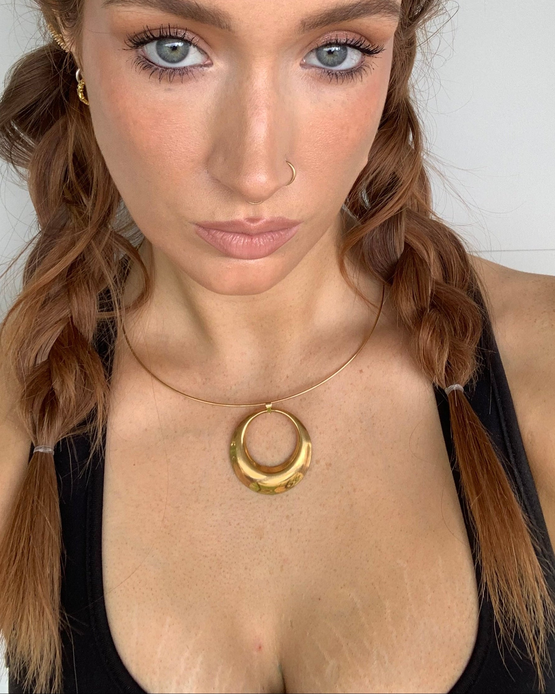 A close-up selfie of a woman with brown hair in a braid, wearing a chunky gold ring necklace and a small gold hoop earring against a white tiled background.