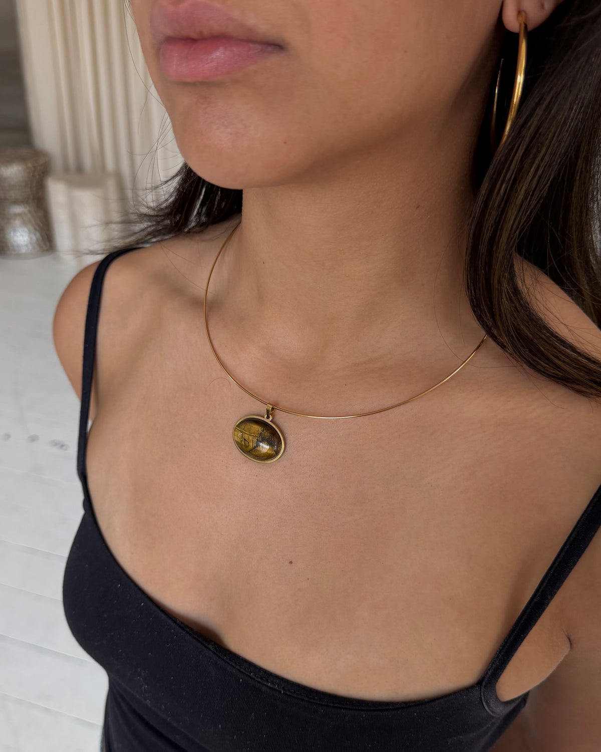 A close-up of a model wearing a delicate gold wire necklace with an oval pendant containing a yellow-green and black pattern, tigers eye stone paired with gold hoop earrings and a black top.
