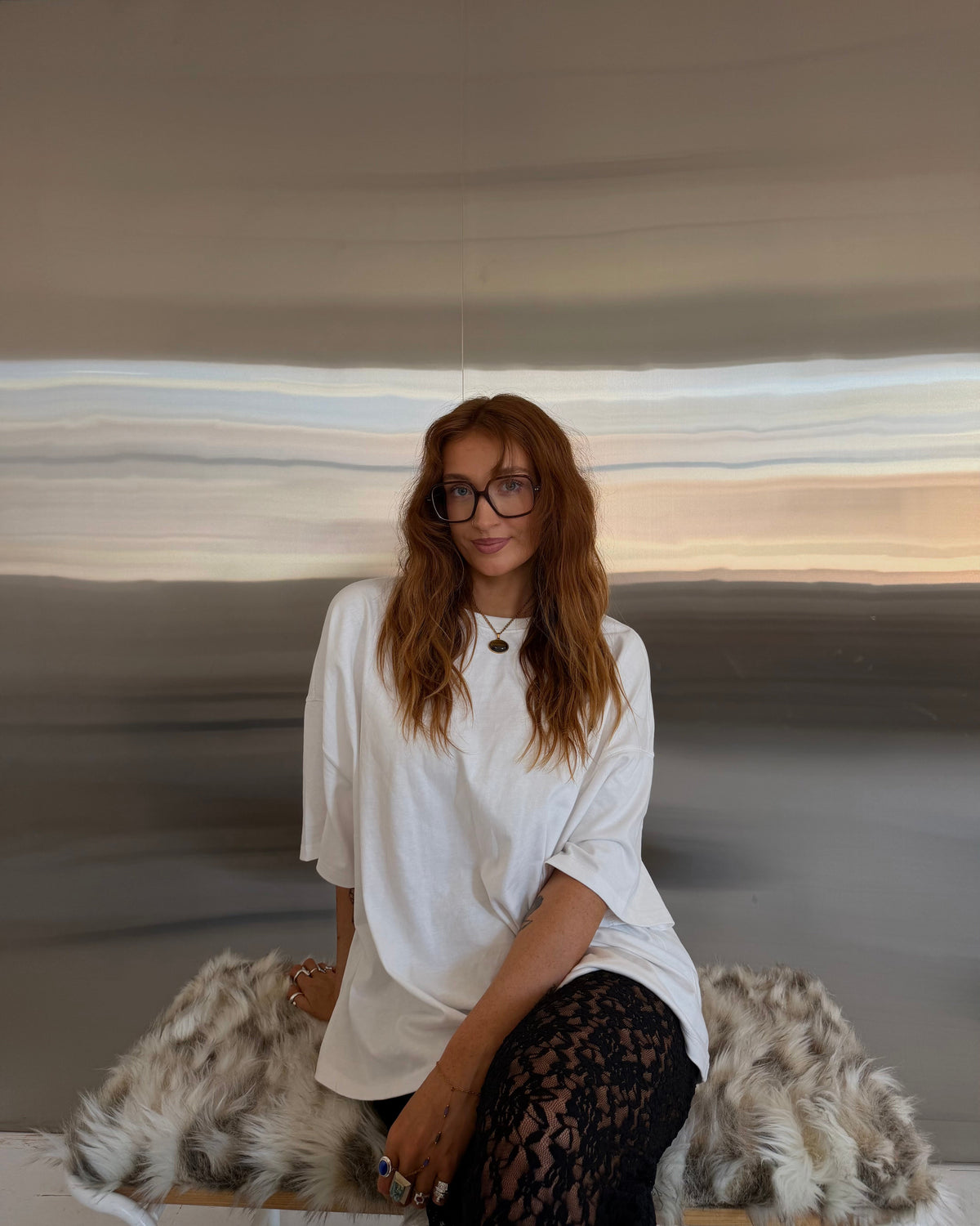 A woman with long auburn hair and glasses sits on a faux fur bench in front of a large abstract landscape painting. She is wearing an oversized white t-shirt and black lace tights.