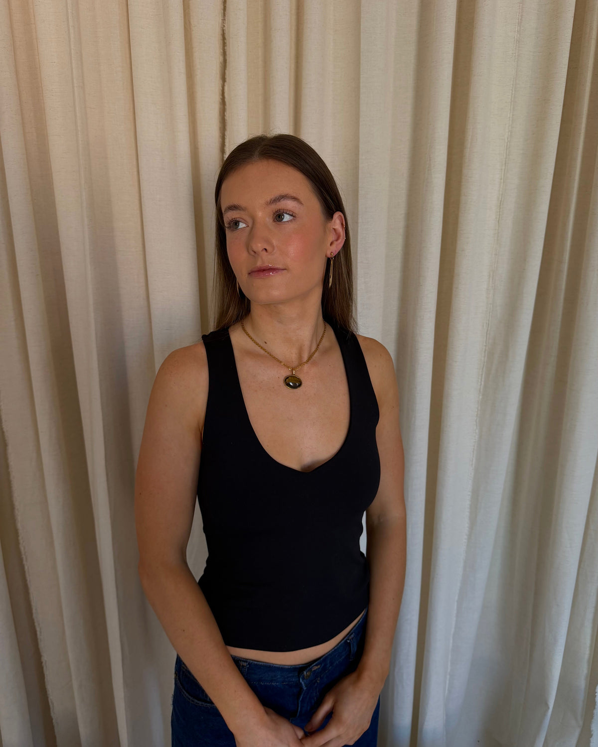 A young woman with light brown hair tied back, wearing a black ribbed tank top and blue jeans, standing in front of a beige curtain backdrop.