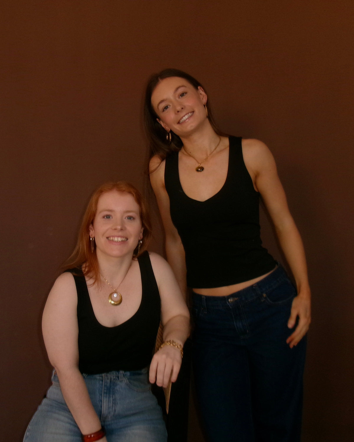 Two young women posing against a solid dark brown wall. The woman on the left is sitting, wearing a black tank top and light blue jeans, while the woman on the right is standing, wearing a black tank top and dark blue jeans.