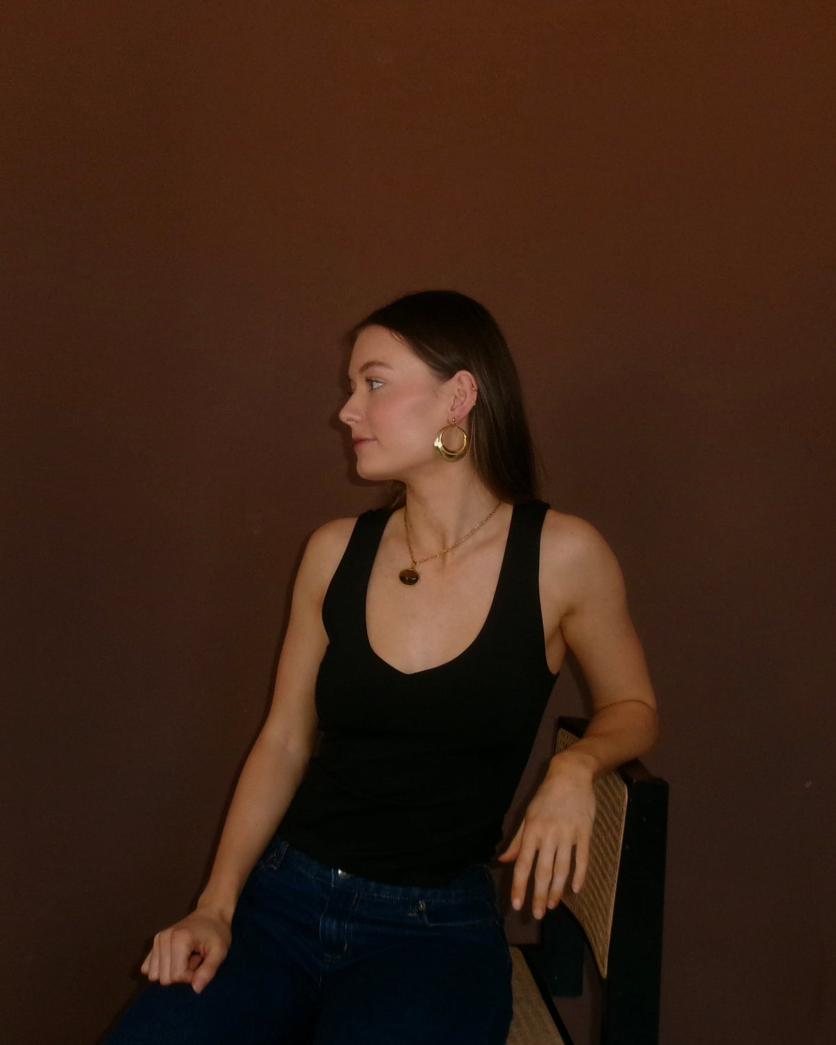 A woman wearing a black ribbed tank top and blue jeans sits against a dark brown wall, looking to the side. She is wearing gold hoop earrings and a necklace.
