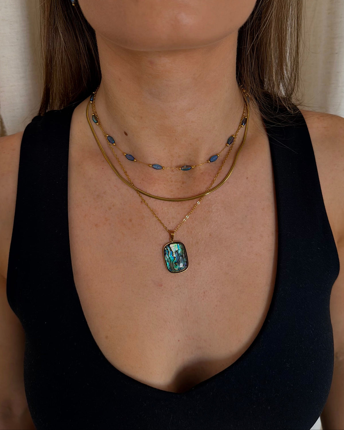 A close-up of a woman wearing layered necklaces, including a gold chain with a green-blue abalone shell pendant and other delicate gold and blue bead chains, paired with a black tank top.