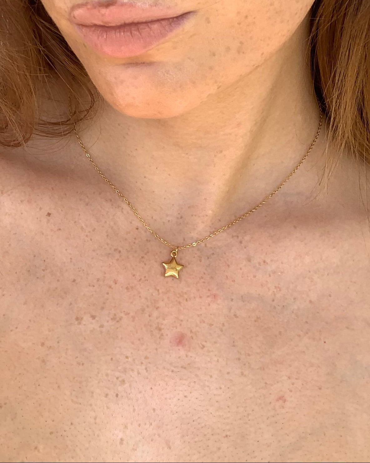 Person with red hair and blue eyes looking directly at the camera against a white tiled wall. wearing a lucky star charm necklace