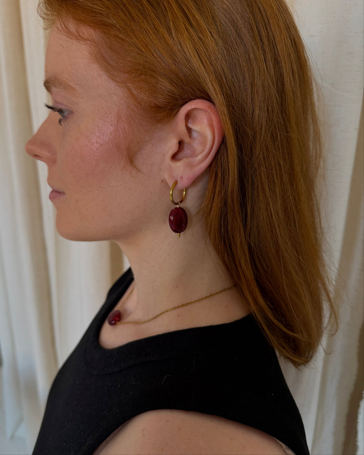 A woman in profile wearing a gold hoop earring with a dark red oval drop pendant, paired with a delicate gold necklace and a black top, set against a soft cream curtain background.