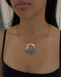 A close-up of a woman wearing a gold wire necklace with a circular pendant made of iridescent mother of pearl shell, paired with a black camisole top.