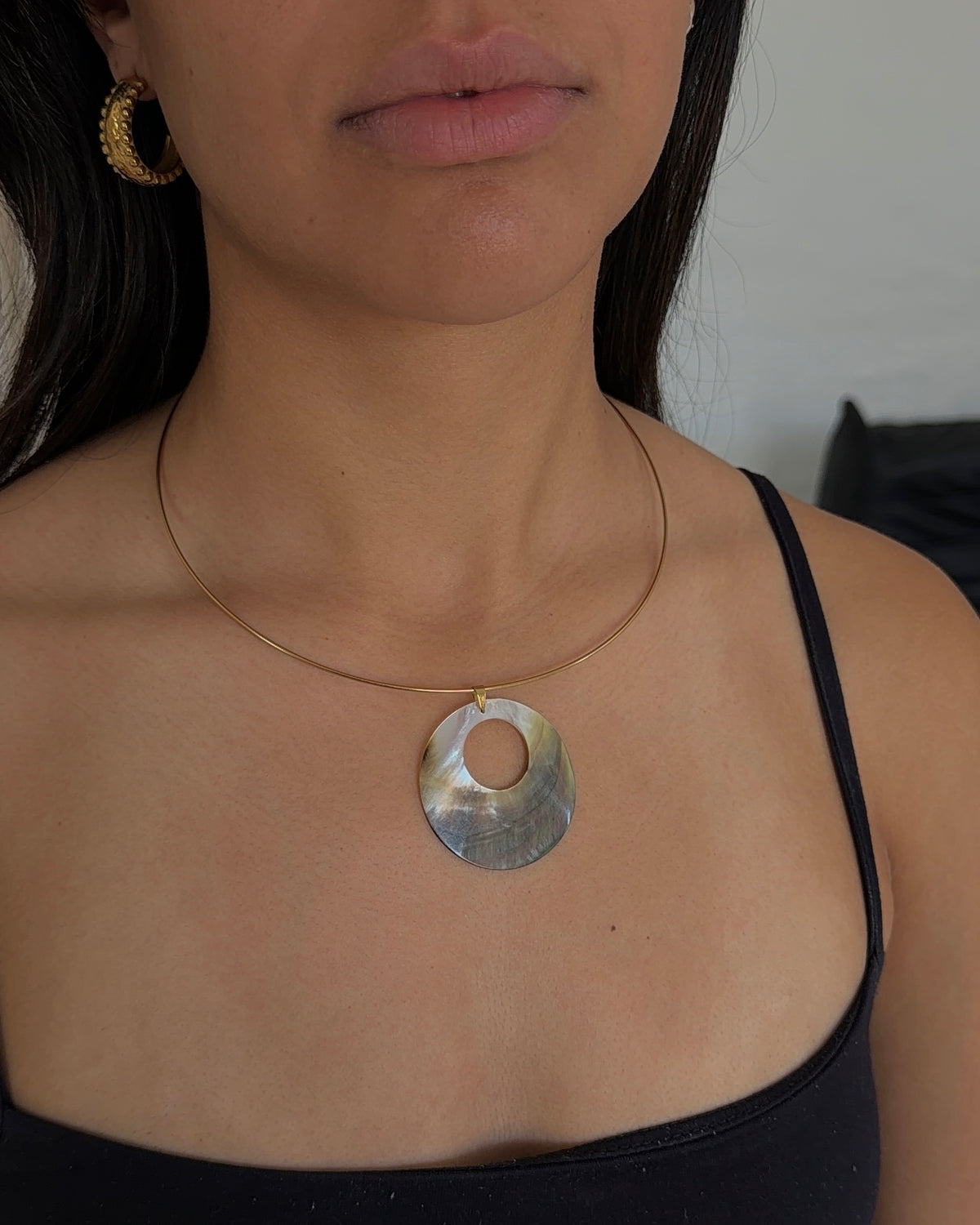 A close-up of a woman wearing a silver wire necklace with a circular pendant made of iridescent mother of pearl shell, paired with a black camisole top.
