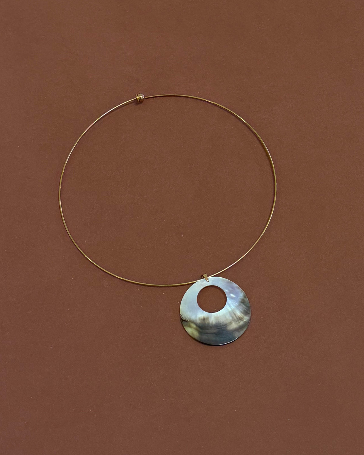 A close-up of a woman wearing a gold wire necklace with a circular pendant made of iridescent mother of pearl shell, paired with a black camisole top.