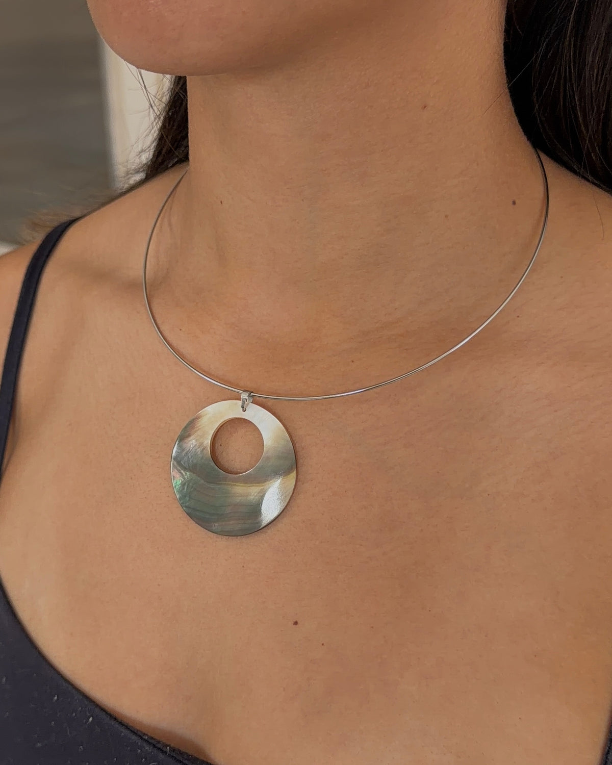 A close-up of a woman wearing a silver wire necklace with a circular pendant made of iridescent mother of pearl shell, paired with a black camisole top.