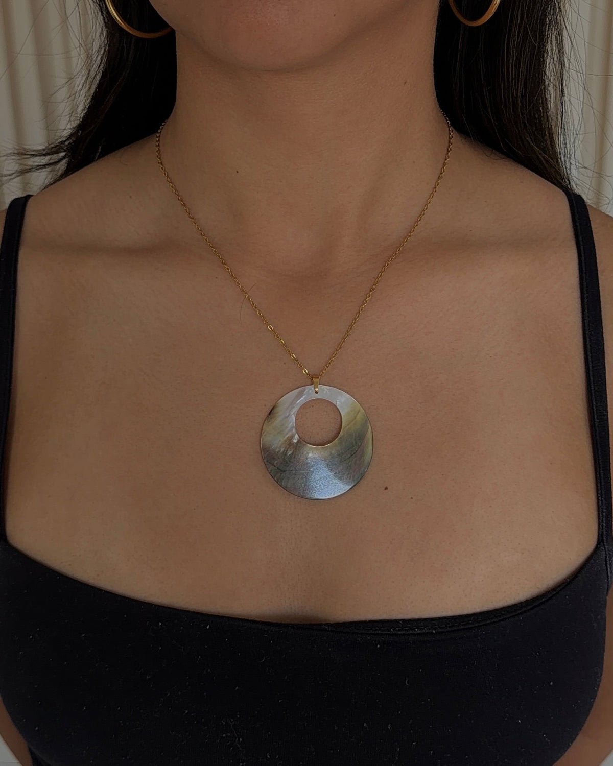 A woman wearing a silver-toned circular pendant necklace with a gold chain, paired with gold hoop earrings and a black camisole top.