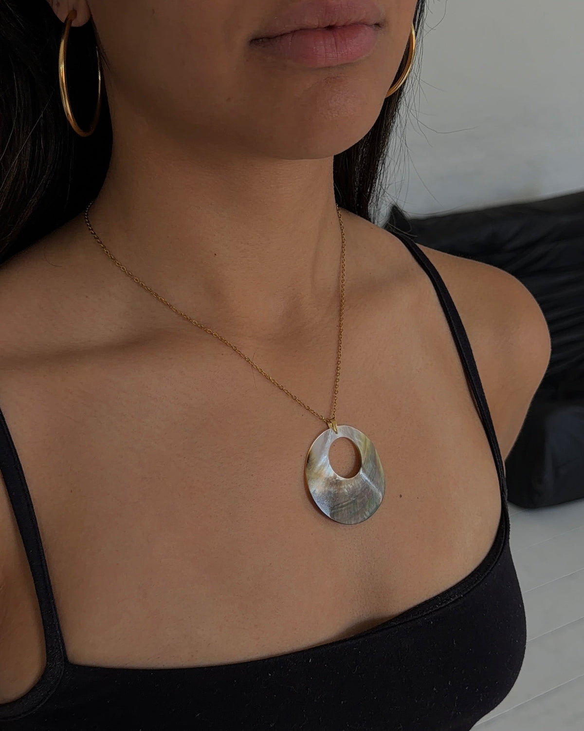 A woman wearing a gold chain necklace with a large, irregular-shaped silver pendant, styled with gold hoop earrings and a black camisole top.