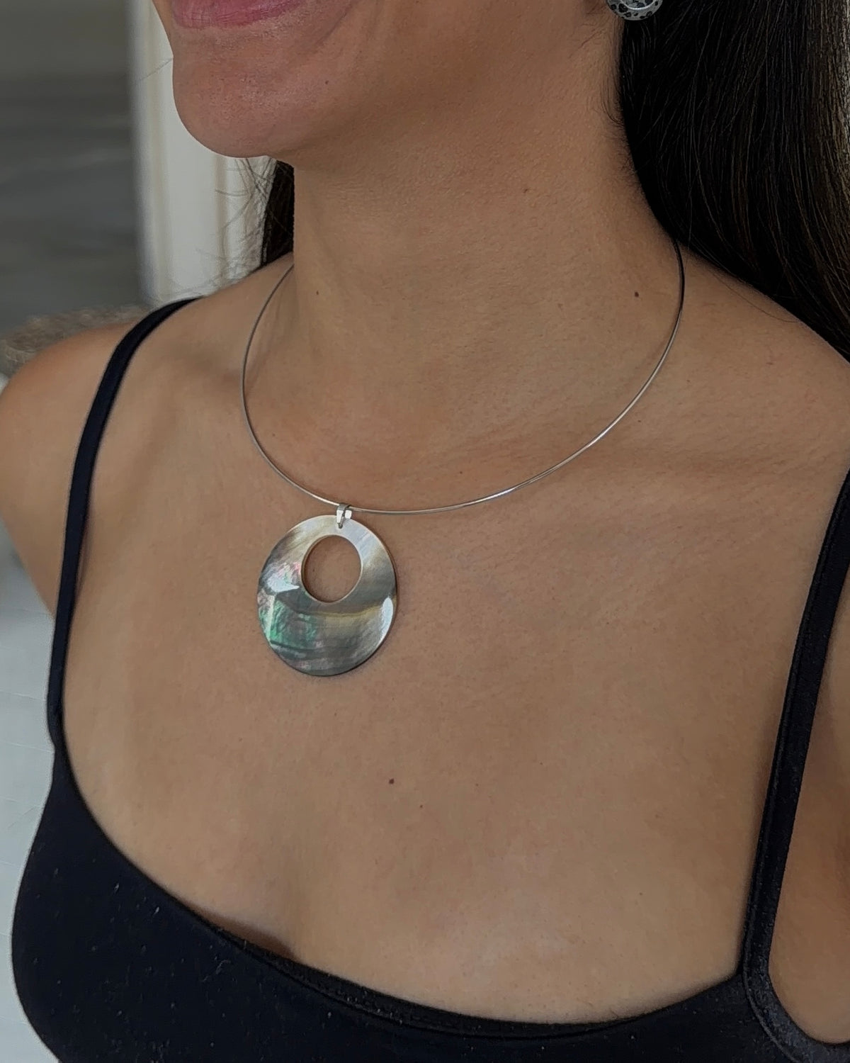 A close-up of a woman wearing a silver wire necklace with a circular pendant made of iridescent mother of pearl shell, paired with a black camisole top.