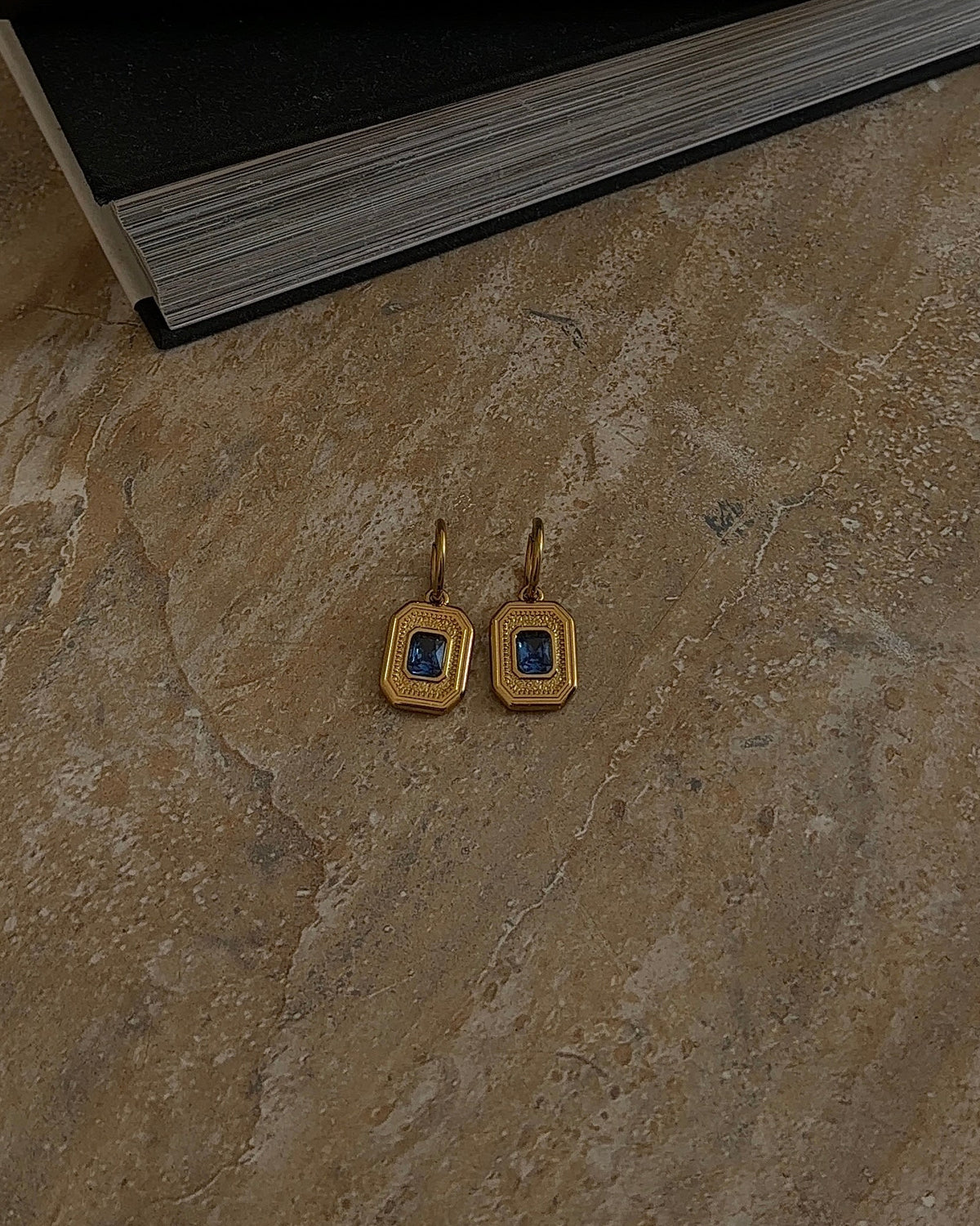 A pair of gold square drop earrings with dark blue square centers, resting on a textured beige stone surface next to a black book.