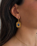 A close-up of a model's ear wearing a gold drop earring with a rectangular blue gemstone, alongside a delicate gold chain necklace.