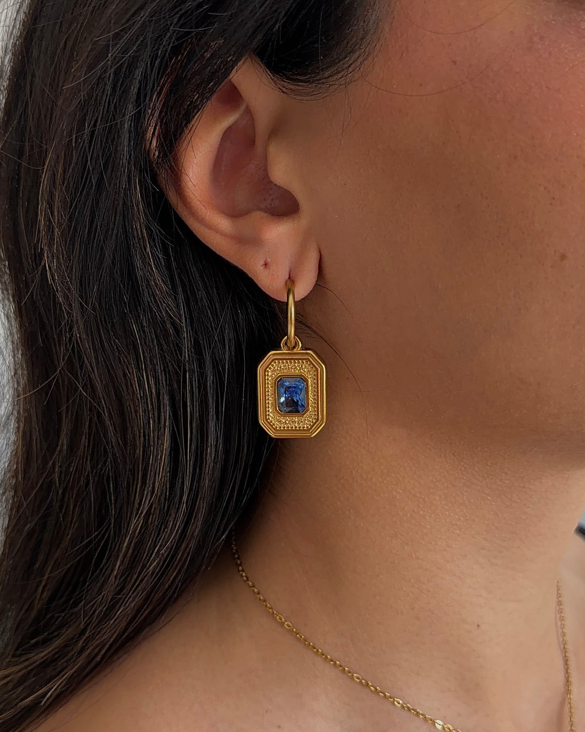 A close-up of a model's ear wearing a gold drop earring with a rectangular blue gemstone, alongside a delicate gold chain necklace.