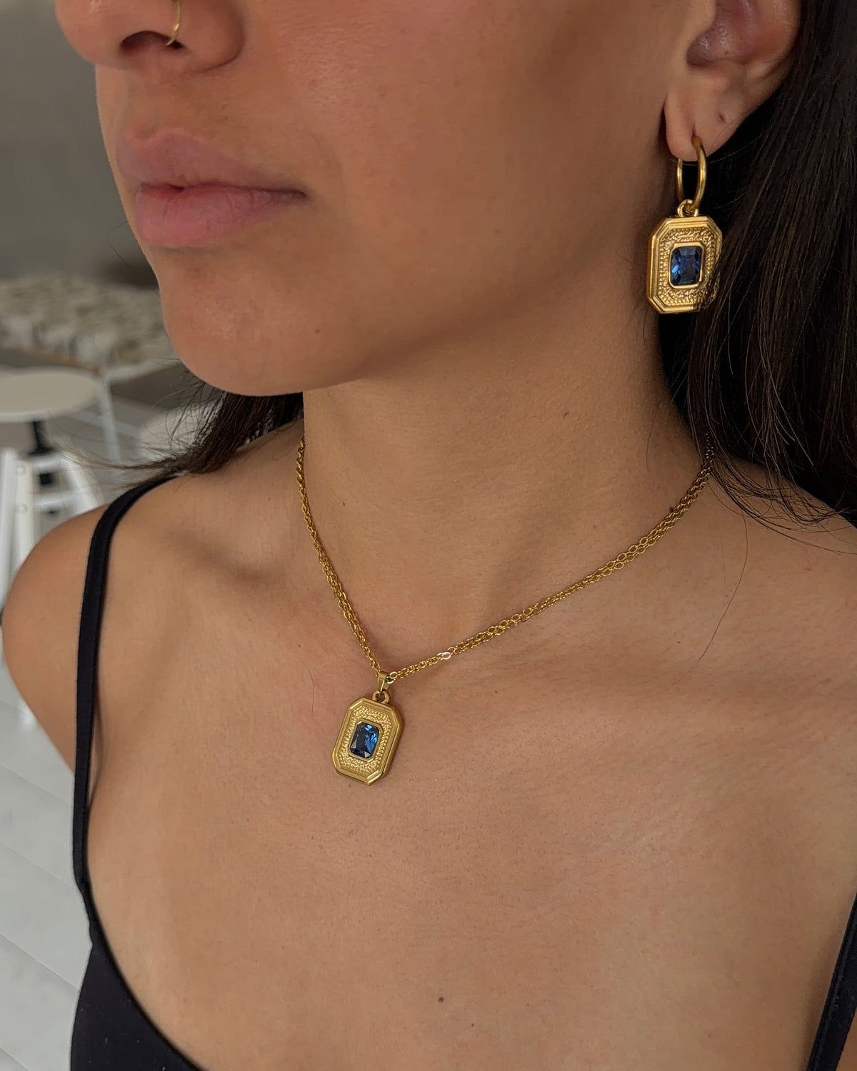 A close-up lifestyle shot of a woman wearing a gold necklace and matching drop earrings, each featuring a rectangular blue gemstone in a gold bezel setting.