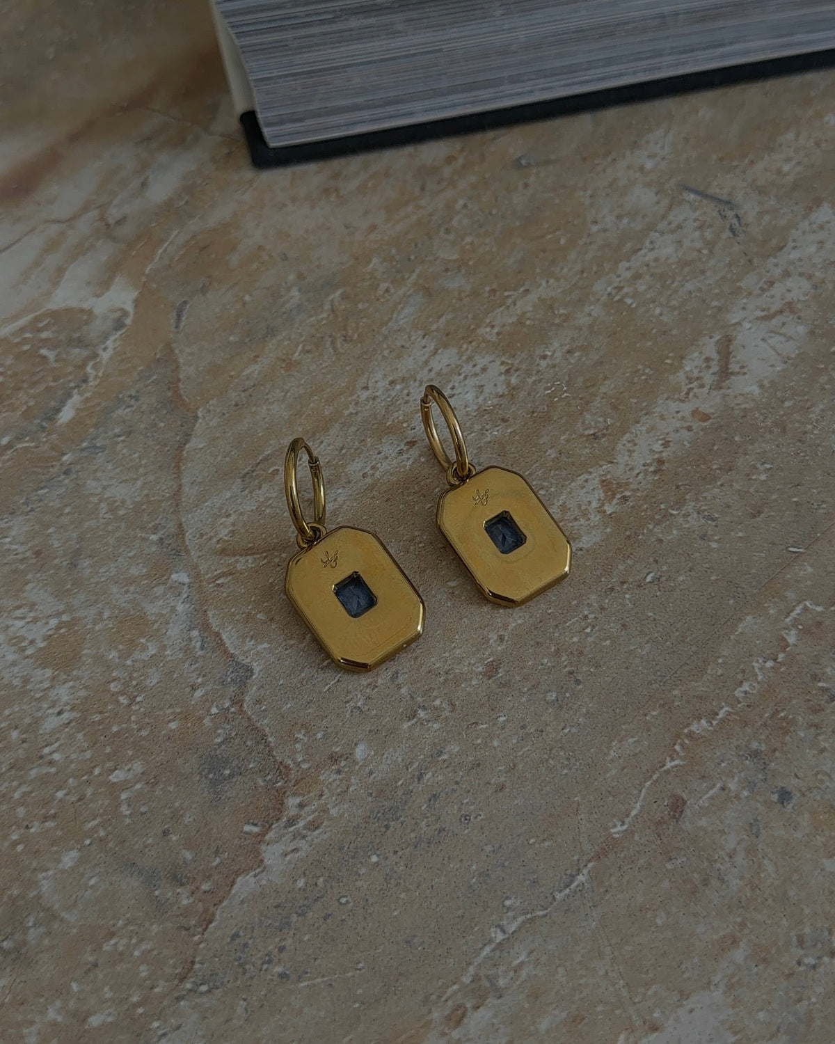 A pair of gold geometric drop earrings with small dark square inlays, resting on a textured beige stone surface next to a dark book.